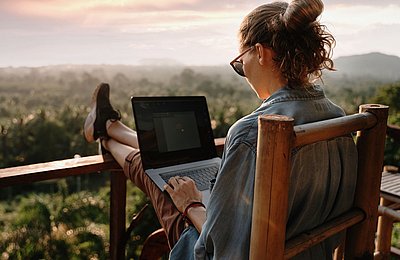 Frau sitzt mit Laptop auf Balkon und arbeitet mit Blick über Baumwipfel bei Sonnenuntergang